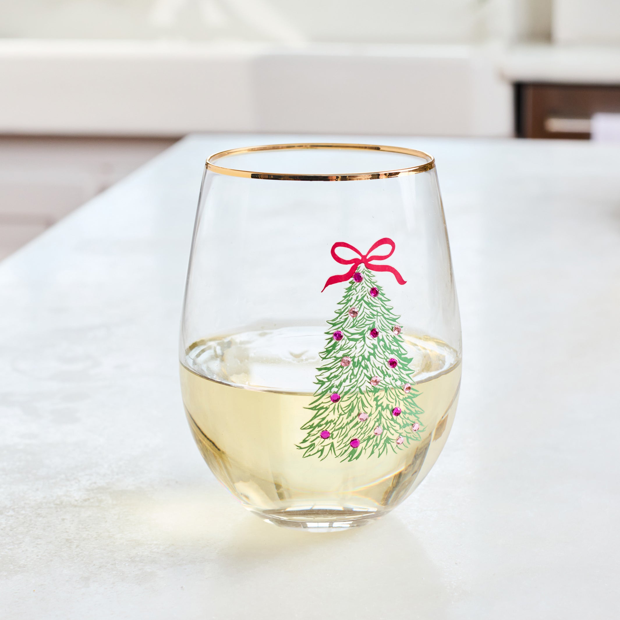 A clear stemless wine glass with a gold rim, filled with white wine and decorated with a green Christmas tree illustration topped with a red bow, set on a light countertop in a bright kitchen.