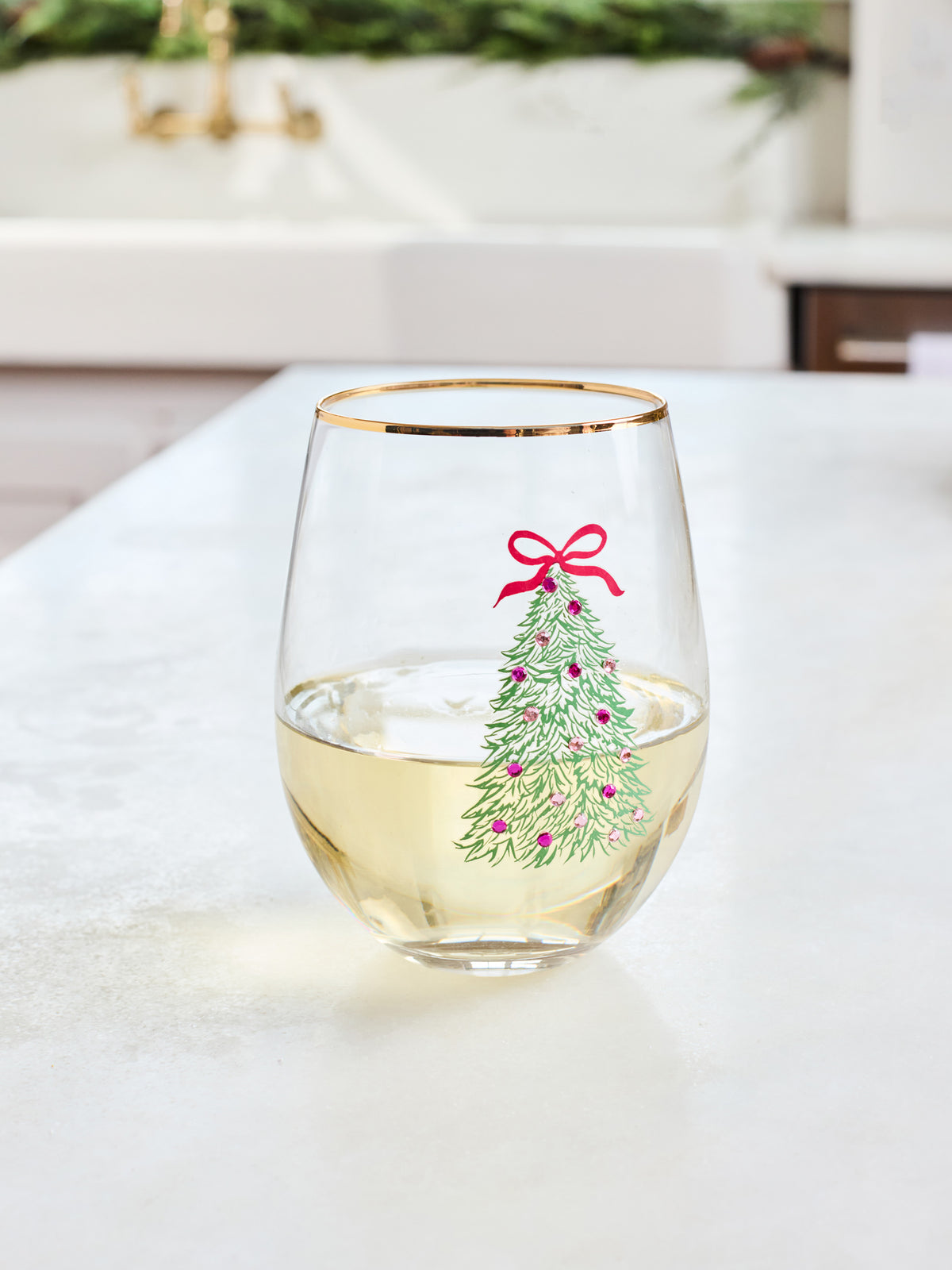 A clear stemless wine glass with a gold rim, filled with white wine and decorated with a green Christmas tree illustration topped with a red bow, set on a light countertop in a bright kitchen.