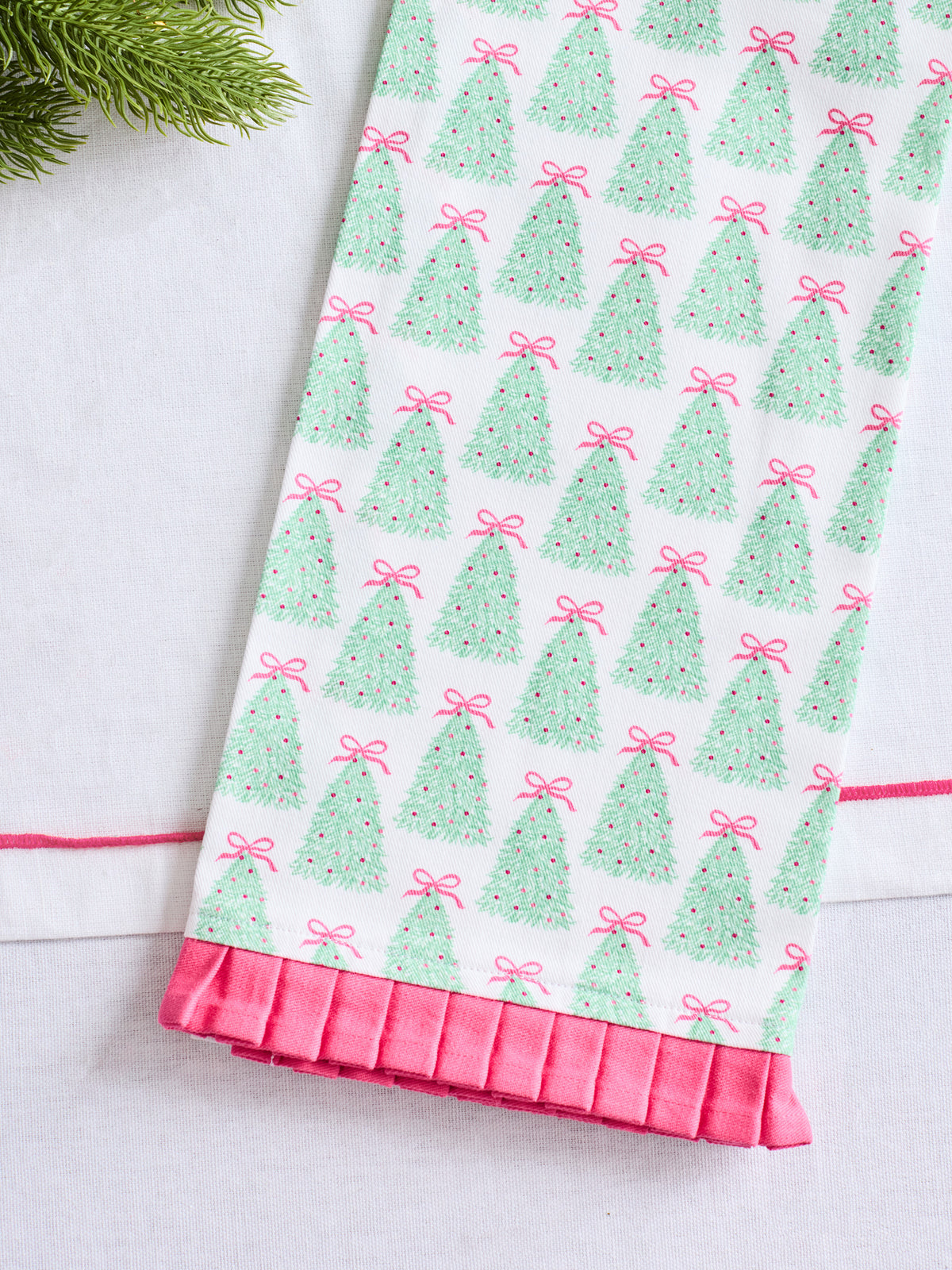 A close-up of a white fabric item, likely a towel or napkin, featuring a repeating pattern of green Christmas trees with pink bows and a pink pleated ruffle trim along the bottom edge.
