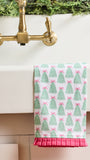 A white kitchen towel with a green Christmas tree and pink bow pattern, draped over the edge of a white sink with a brass faucet and greenery in the background.