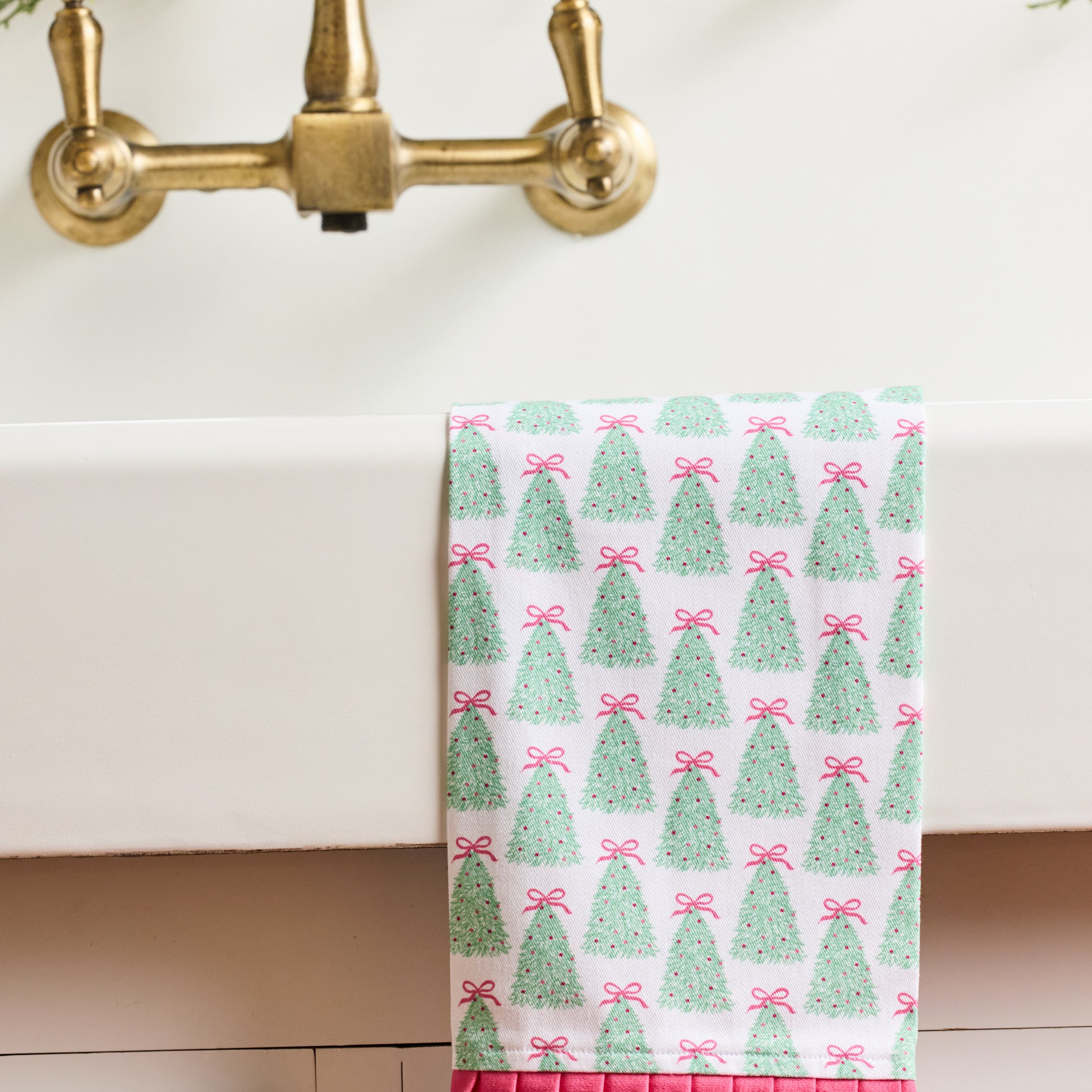 A white kitchen towel with a green Christmas tree and pink bow pattern, draped over the edge of a white sink with a brass faucet and greenery in the background.