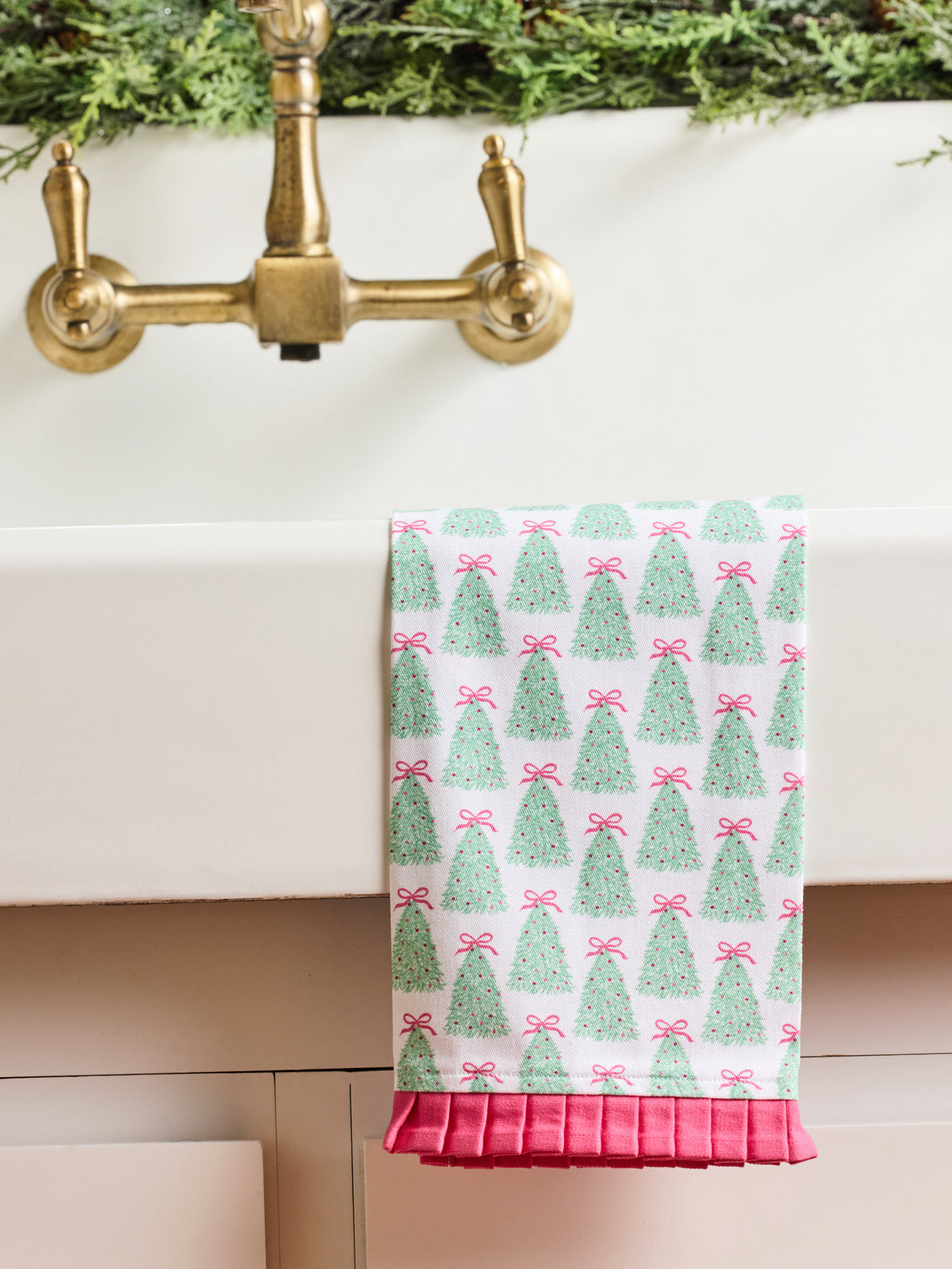 A white kitchen towel with a green Christmas tree and pink bow pattern, draped over the edge of a white sink with a brass faucet and greenery in the background.