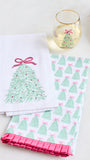 Two white kitchen towels with Christmas tree designs, one featuring a single green tree with a pink bow and the other with a repeating green and pink tree pattern, styled on a white surface with a wine glass and gold tray.