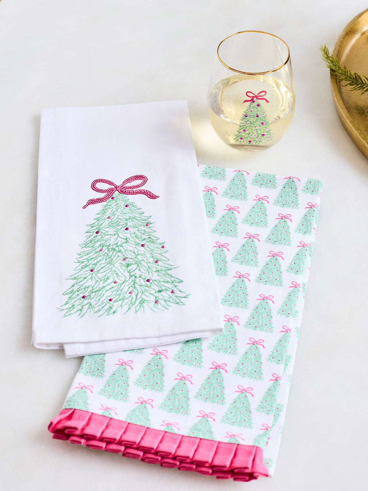 Two white kitchen towels with Christmas tree designs, one featuring a single green tree with a pink bow and the other with a repeating green and pink tree pattern, styled on a white surface with a wine glass and gold tray.