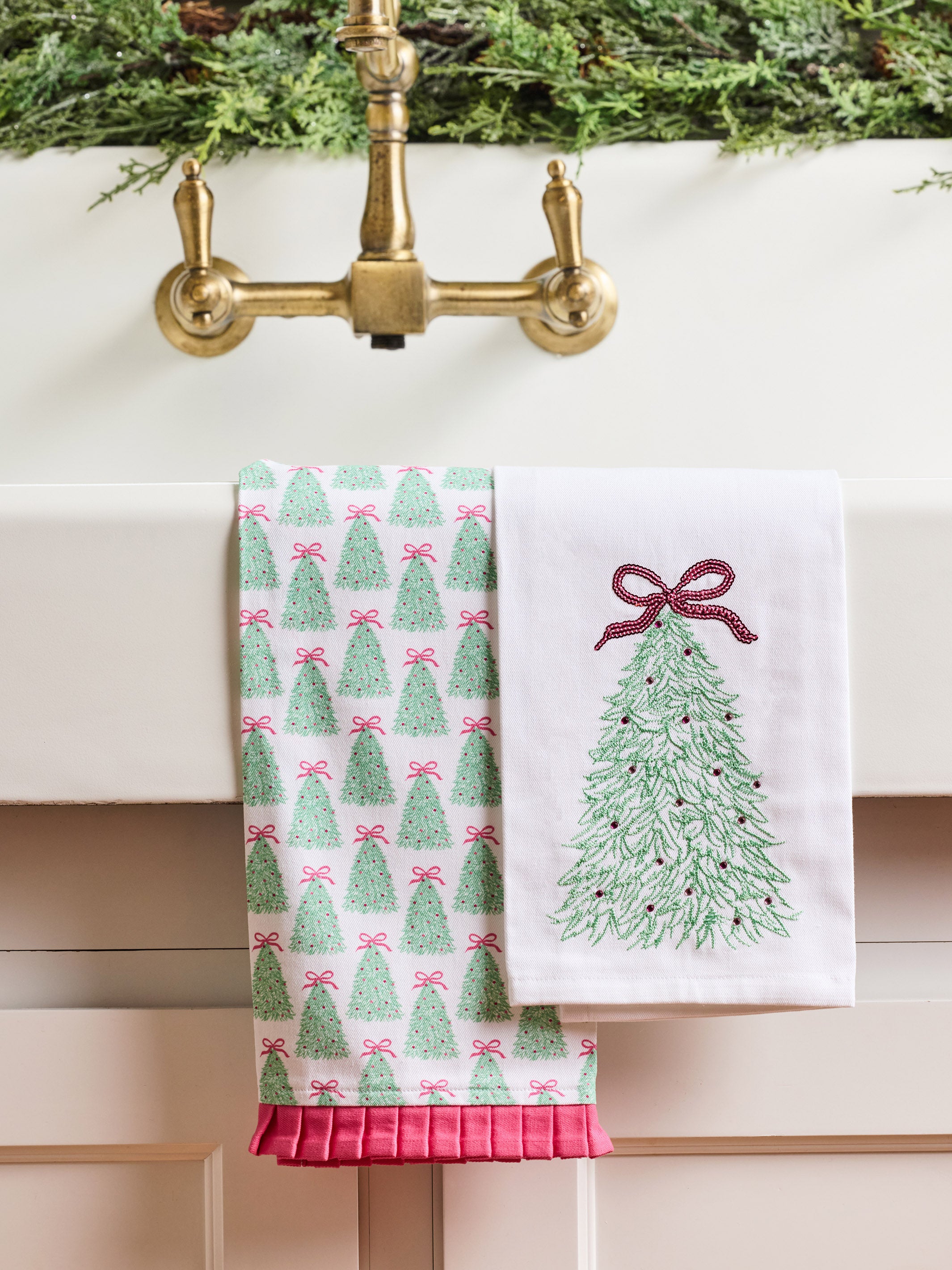 Two white kitchen towels hanging over a brass faucet. One towel has a repeating green Christmas tree pattern with pink bows, and the other has a single large green Christmas tree design with a pink bow on the right side.
