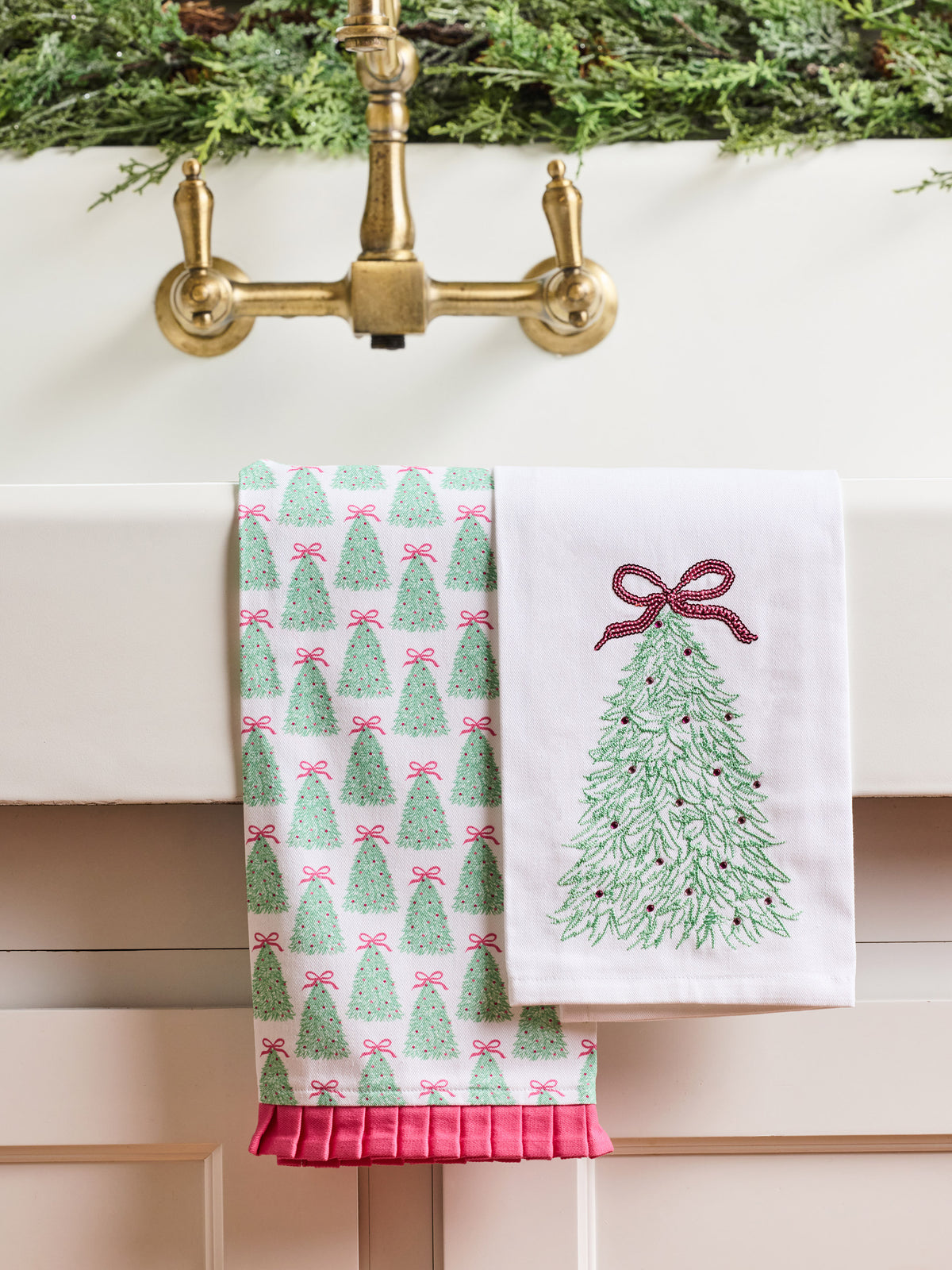 Two white kitchen towels hanging over a brass faucet. One towel has a repeating green Christmas tree pattern with pink bows, and the other has a single large green Christmas tree design with a pink bow on the right side.
