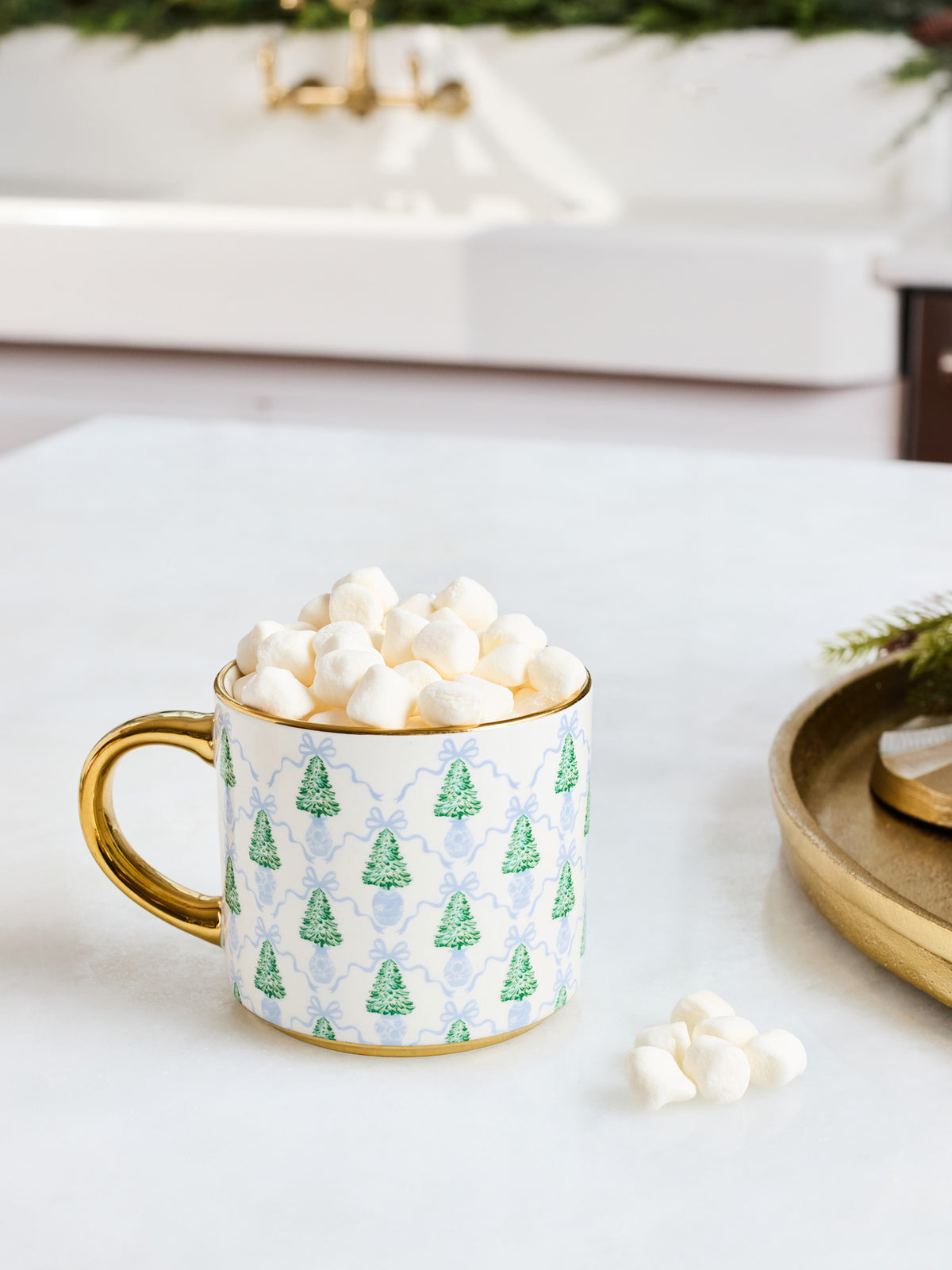 A white ceramic mug with a gold handle and rim, decorated with a green Christmas tree and blue damask pattern, filled with mini marshmallows on a white countertop.
