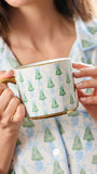 A person in light blue pajamas holds a white ceramic mug with a green Christmas tree pattern and gold trim.