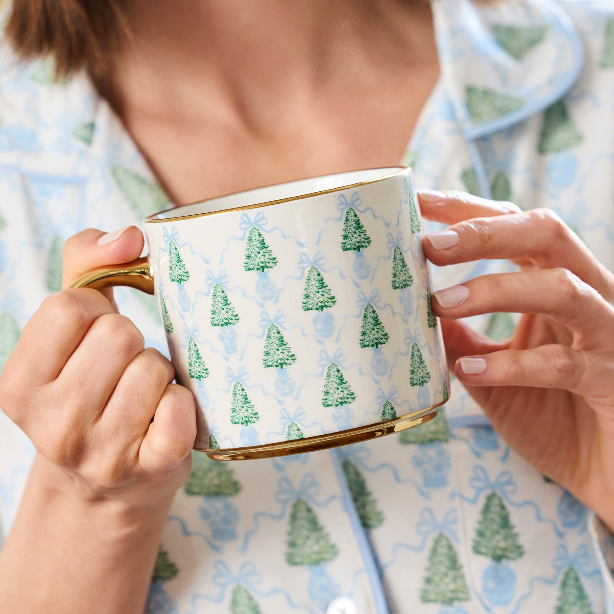A person in light blue pajamas holds a white ceramic mug with a green Christmas tree pattern and gold trim.