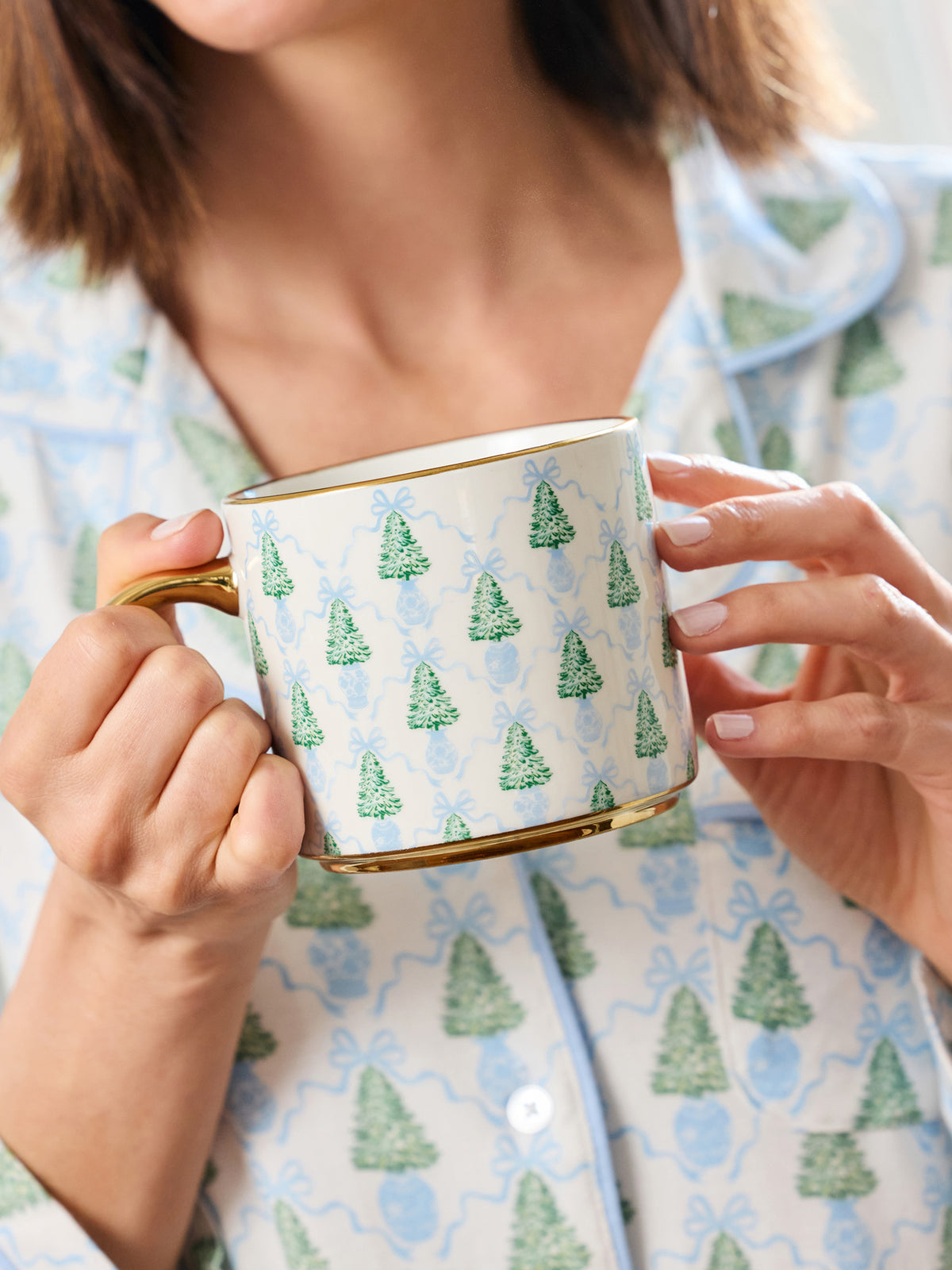 A person in light blue pajamas holds a white ceramic mug with a green Christmas tree pattern and gold trim.
