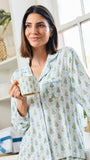 A woman wearing a light blue and white long-sleeve pajama set with a green pine tree pattern, holding a matching mug in a bright, cozy room.