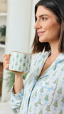 A woman in light blue pajamas with a green Christmas tree pattern holds a matching mug with a gold rim, set against a bright, softly blurred indoor background.