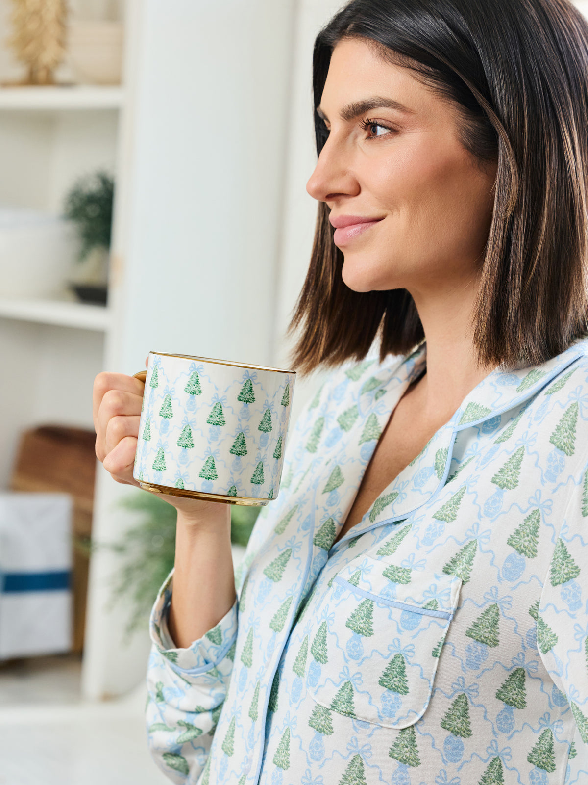 A woman in light blue pajamas with a green Christmas tree pattern holds a matching mug with a gold rim, set against a bright, softly blurred indoor background.