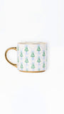 A white ceramic mug with a gold handle and base trim, decorated with a repeating pattern of green Christmas trees and light blue damask motifs.