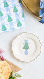 A scalloped white ceramic plate with a gold rim and a blue-and-green Christmas tree illustration, styled on a white surface with bread, sliced red onion, a patterned napkin, and a gold tray.