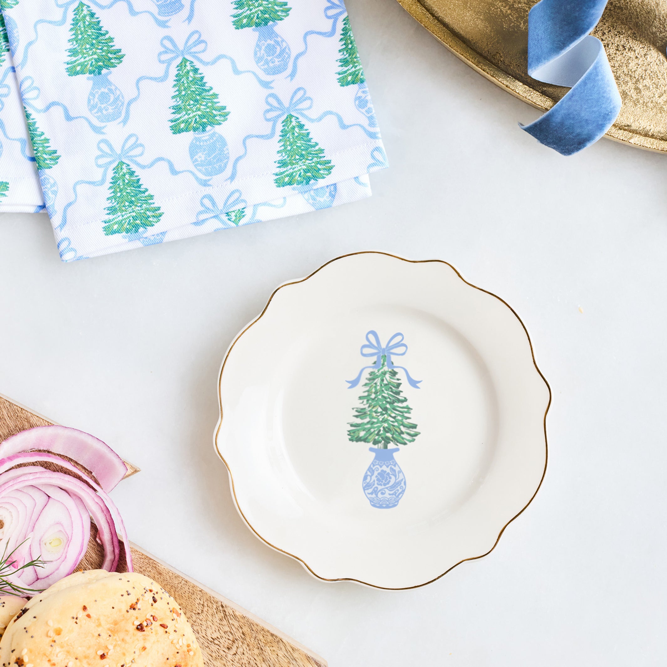 A scalloped white ceramic plate with a gold rim and a blue-and-green Christmas tree illustration, styled on a white surface with bread, sliced red onion, a patterned napkin, and a gold tray.