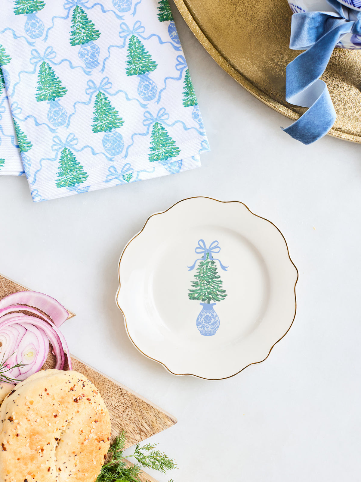 A scalloped white ceramic plate with a gold rim and a blue-and-green Christmas tree illustration, styled on a white surface with bread, sliced red onion, a patterned napkin, and a gold tray.