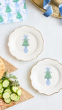 Two scalloped white ceramic plates with gold trim, each decorated with a blue and green Christmas tree motif, arranged on a white tabletop with food props including sliced cucumbers, bread, and a patterned napkin.
