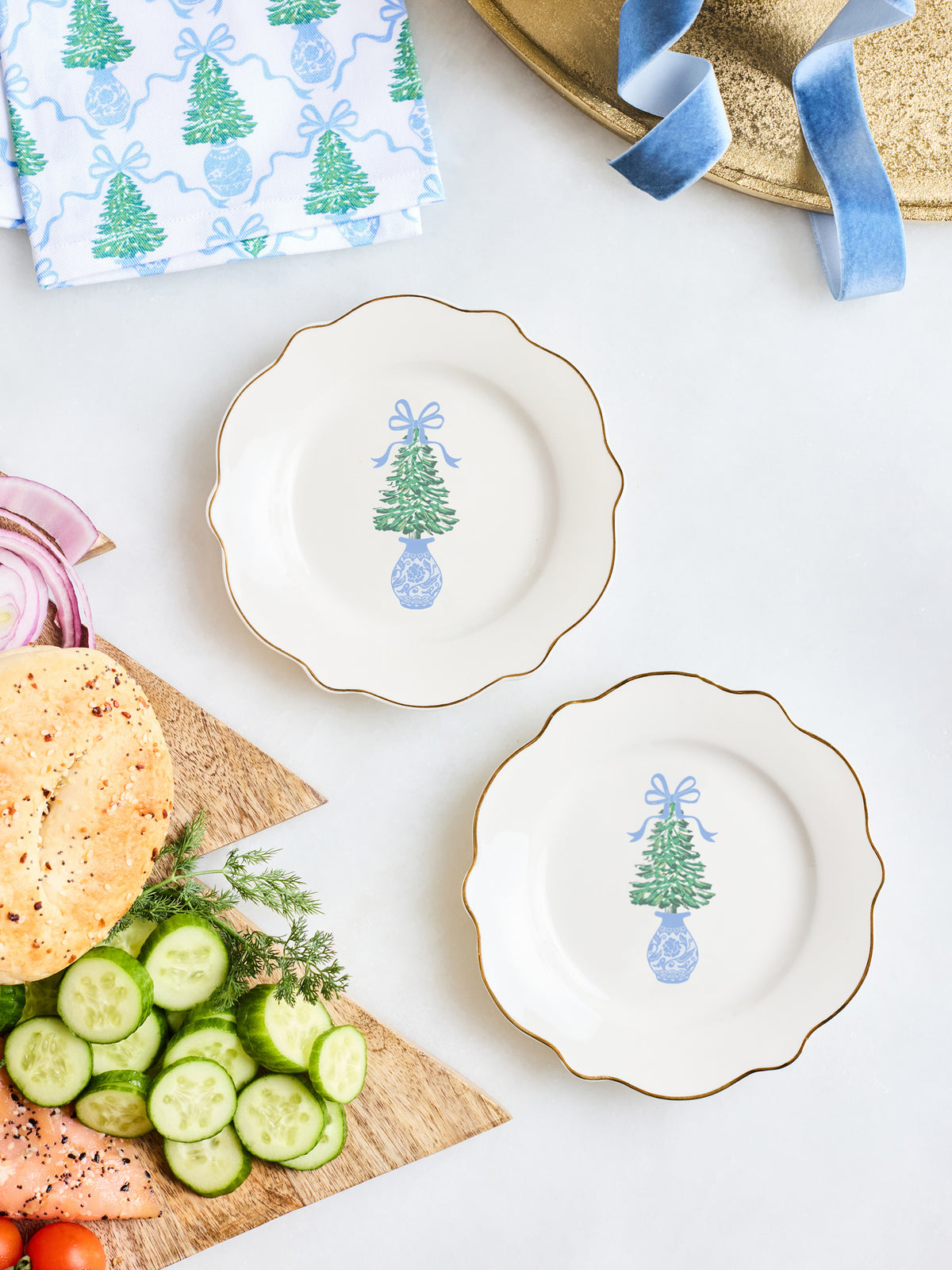 Two scalloped white ceramic plates with gold trim, each decorated with a blue and green Christmas tree motif, arranged on a white tabletop with food props including sliced cucumbers, bread, and a patterned napkin.