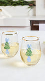 Two clear stemless wine glasses with gold rims, filled with white wine, decorated with a green Christmas tree, blue bow, and blue ornament design, set on a white countertop in a bright kitchen.