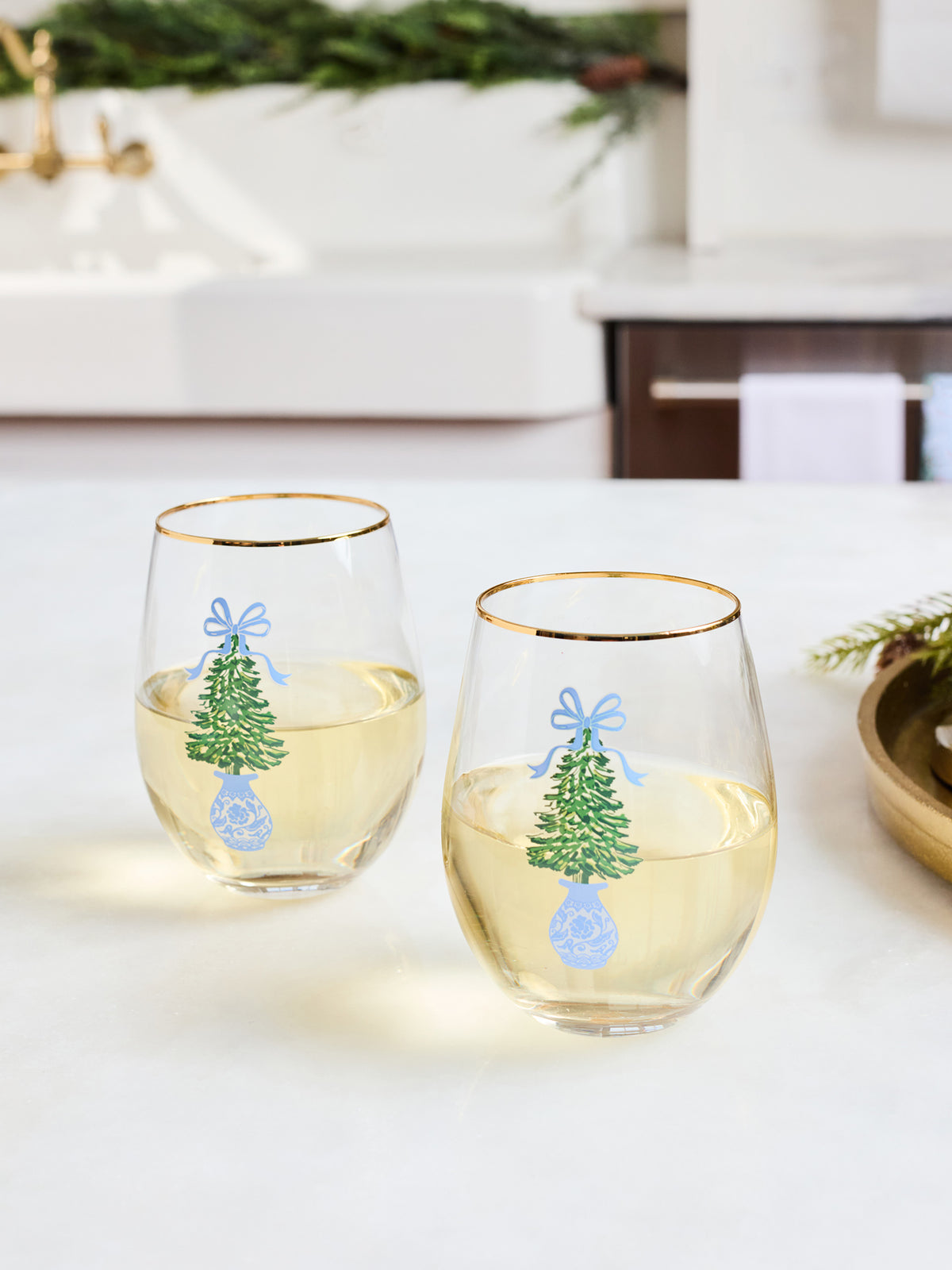 Two clear stemless wine glasses with gold rims, filled with white wine, decorated with a green Christmas tree, blue bow, and blue ornament design, set on a white countertop in a bright kitchen.