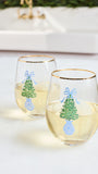 Two clear stemless wine glasses with gold rims, decorated with a green Christmas tree, blue bow, and blue ornament design, filled with white wine on a light tabletop.