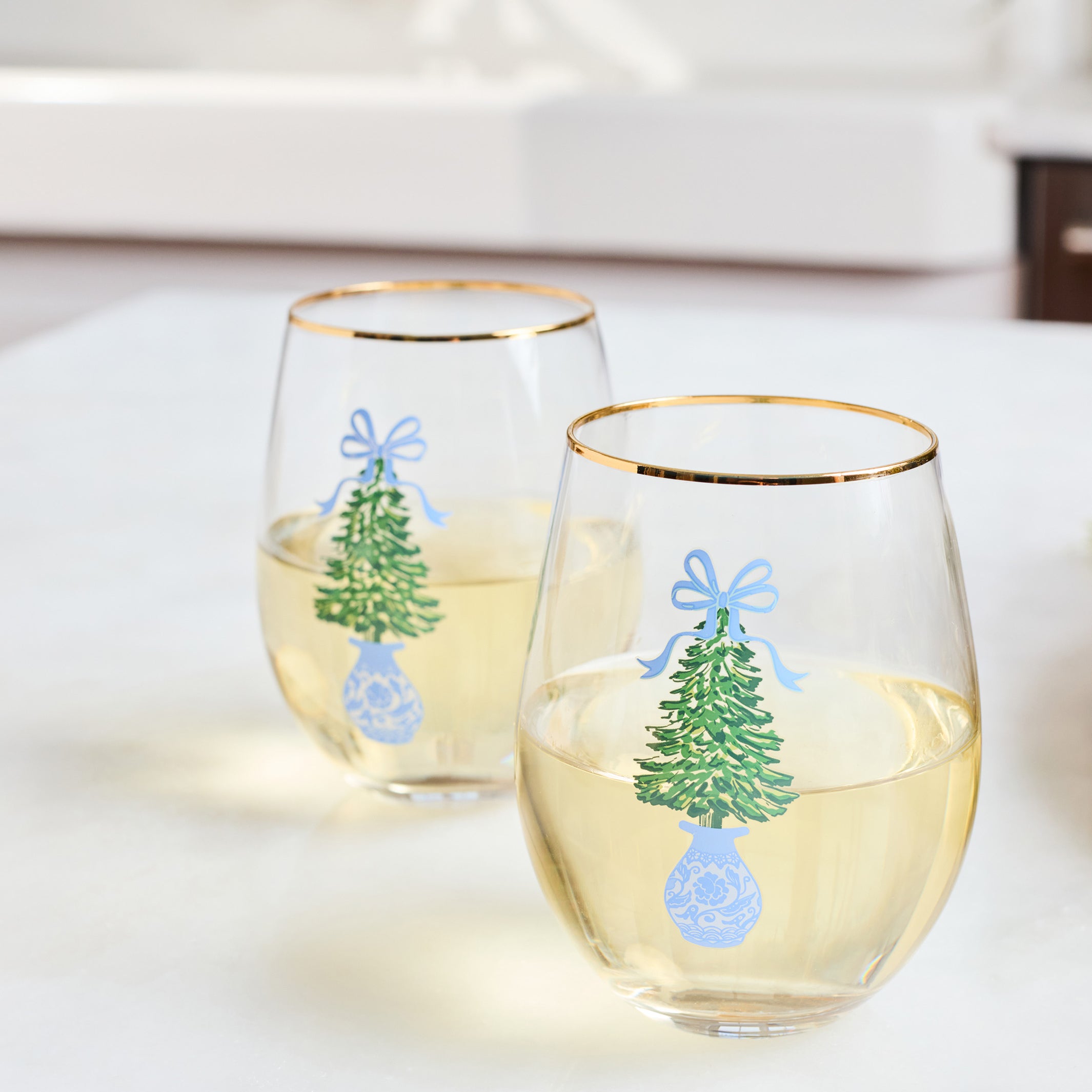 Two clear stemless wine glasses with gold rims, decorated with a green Christmas tree, blue bow, and blue ornament design, filled with white wine on a light tabletop.