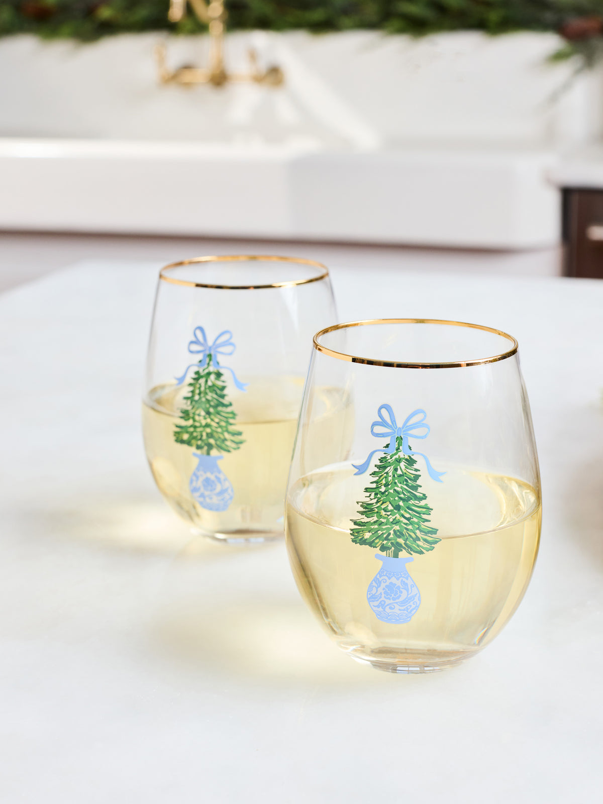 Two clear stemless wine glasses with gold rims, decorated with a green Christmas tree, blue bow, and blue ornament design, filled with white wine on a light tabletop.