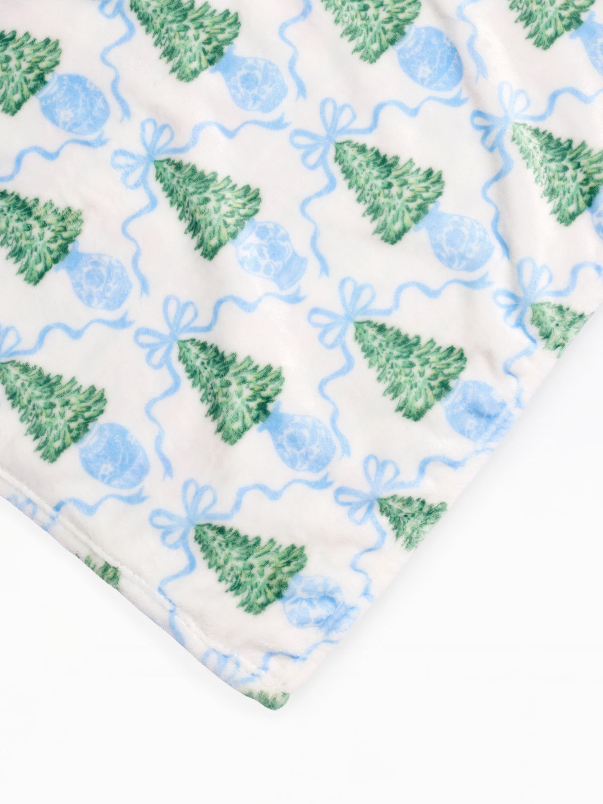 A close-up of a soft fabric product, likely a blanket or throw, featuring a repeating pattern of green Christmas trees, blue ornaments, and wavy blue lines on an off-white background.