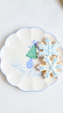 A scalloped white ceramic plate with a blue rim, decorated with a green Christmas tree and the words 'Merry Christmas' in blue script. Two decorated sugar cookies, one snowflake-shaped in light blue icing and one round with blue icing, rest on the plate against a light background.