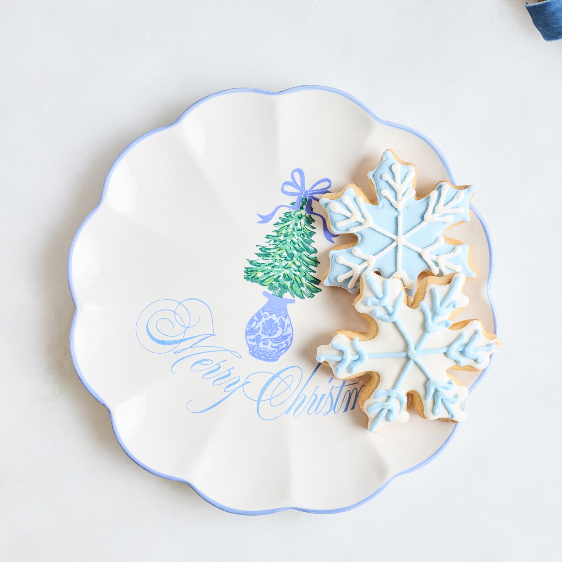 A scalloped white ceramic plate with a blue rim, decorated with a green Christmas tree and the words 'Merry Christmas' in blue script. Two decorated sugar cookies, one snowflake-shaped in light blue icing and one round with blue icing, rest on the plate against a light background.