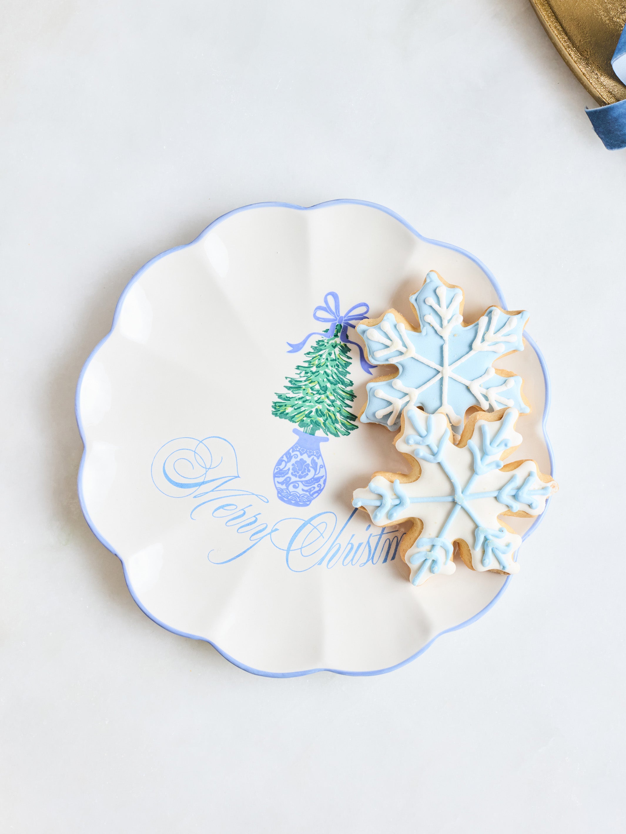 A scalloped white ceramic plate with a blue rim, decorated with a green Christmas tree and the words 'Merry Christmas' in blue script. Two decorated sugar cookies, one snowflake-shaped in light blue icing and one round with blue icing, rest on the plate against a light background.