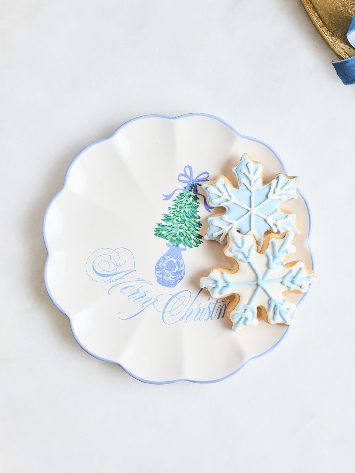 A scalloped white ceramic plate with a blue rim, decorated with a green Christmas tree and the words 'Merry Christmas' in blue script. Two decorated sugar cookies, one snowflake-shaped in light blue icing and one round with blue icing, rest on the plate against a light background.