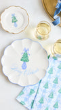 A styled holiday table setting featuring a large scalloped white plate with a blue rim and a green Christmas tree illustration, accompanied by a smaller matching plate, patterned napkins, and two glasses of white wine on a light background.