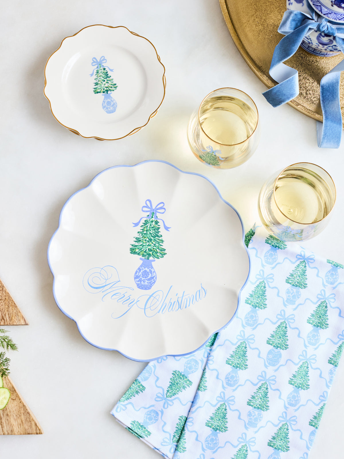A styled holiday table setting featuring a large scalloped white plate with a blue rim and a green Christmas tree illustration, accompanied by a smaller matching plate, patterned napkins, and two glasses of white wine on a light background.
