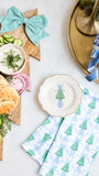 A styled holiday tabletop scene with a wooden star board holding sliced cucumbers, bagels, onion, and a small bowl of dill sauce, accented by a mint green striped bow. A folded white napkin with blue Christmas tree and gift patterns sits in front, alongside a white plate with a blue tree illustration and a blue-and-white ceramic bowl on a gold tray.