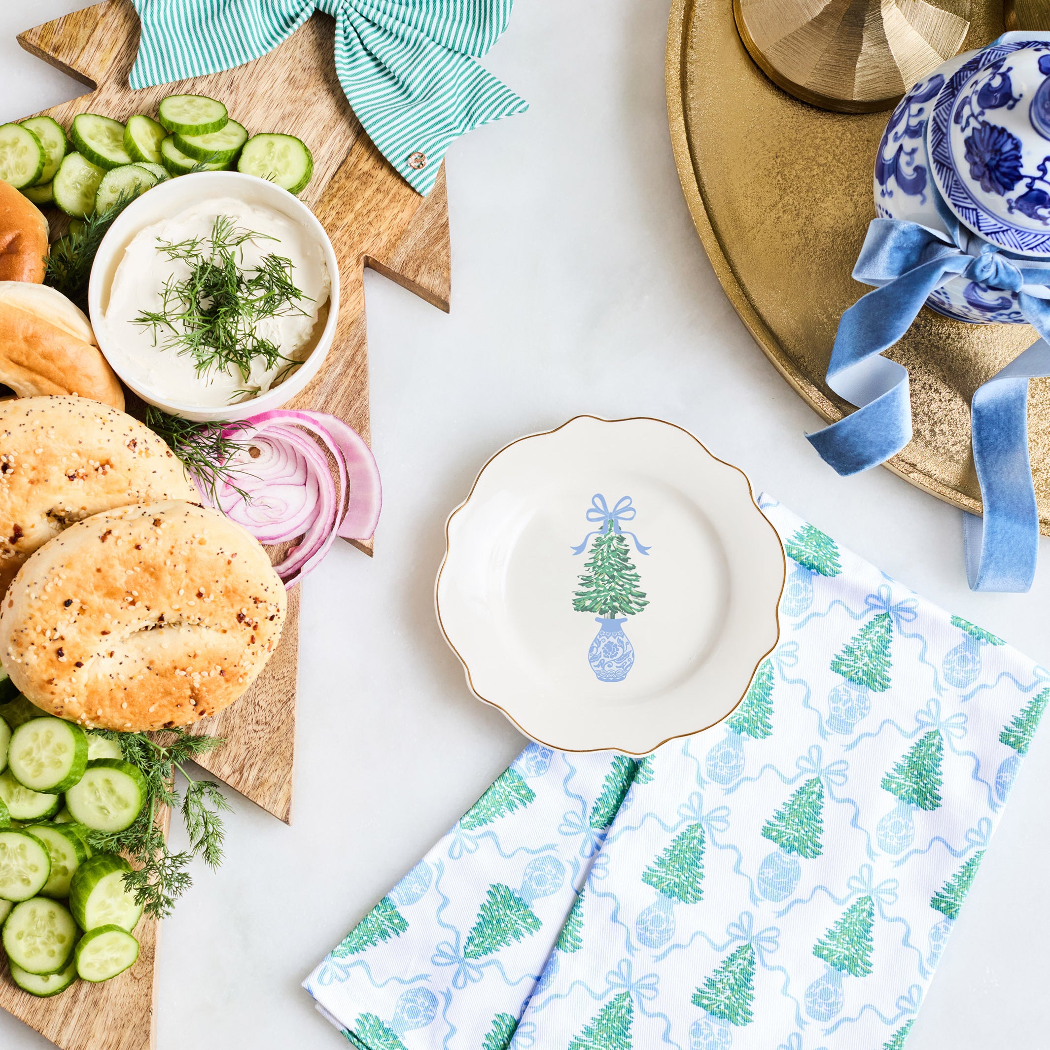 A styled holiday tabletop scene with a wooden star board holding sliced cucumbers, bagels, onion, and a small bowl of dill sauce, accented by a mint green striped bow. A folded white napkin with blue Christmas tree and gift patterns sits in front, alongside a white plate with a blue tree illustration and a blue-and-white ceramic bowl on a gold tray.