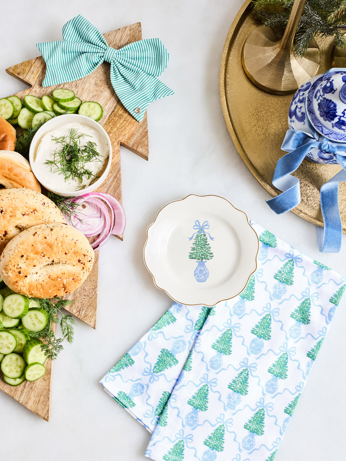 A styled holiday tabletop scene with a wooden star board holding sliced cucumbers, bagels, onion, and a small bowl of dill sauce, accented by a mint green striped bow. A folded white napkin with blue Christmas tree and gift patterns sits in front, alongside a white plate with a blue tree illustration and a blue-and-white ceramic bowl on a gold tray.