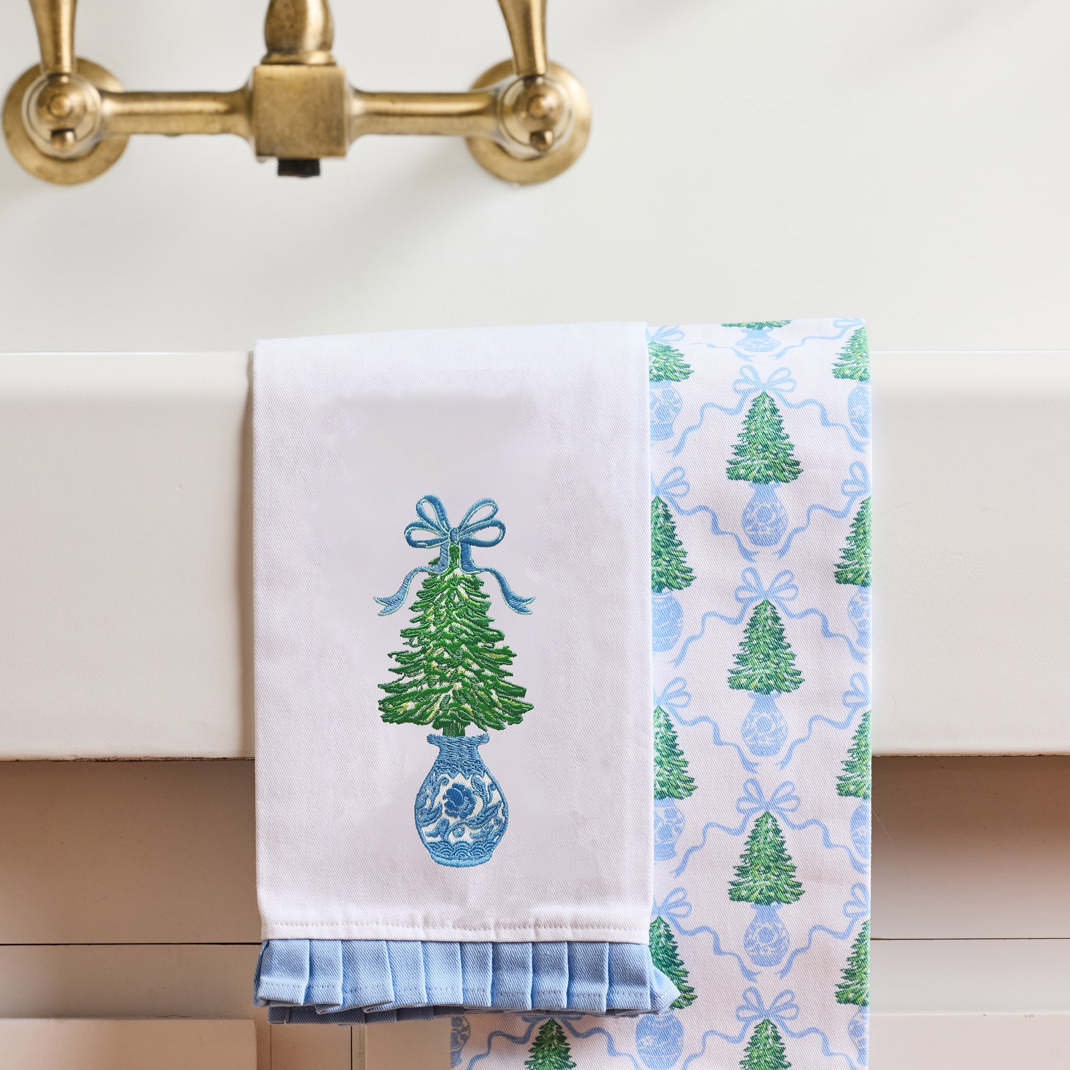 Two decorative kitchen towels hanging over a white sink. One towel is white with an embroidered green Christmas tree in a blue vase with a blue bow.