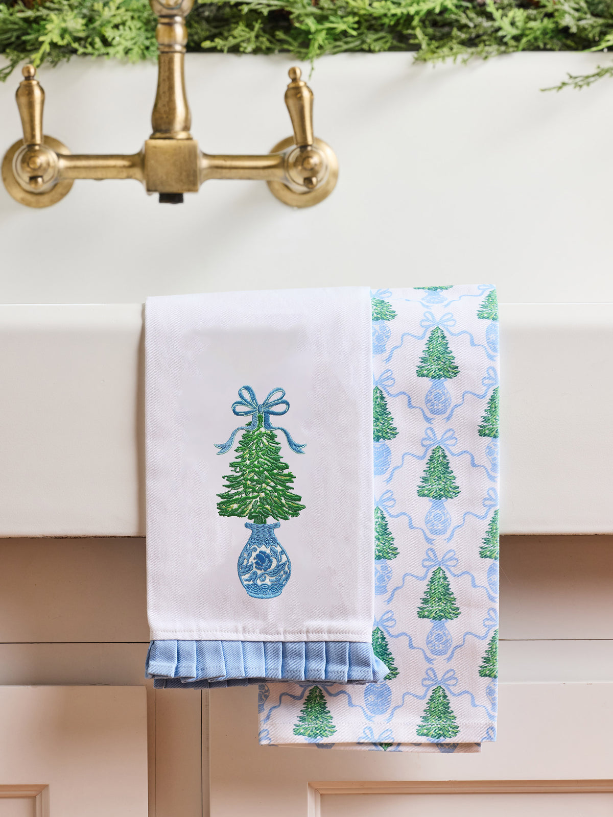 Two decorative kitchen towels hanging over a white sink. One towel is white with an embroidered green Christmas tree in a blue vase with a blue bow.