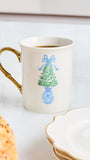 A white ceramic mug with a gold handle, filled with coffee, decorated with a blue and green Christmas tree in a blue vase. The mug is set on a light marble surface next to a plate and a pastry.