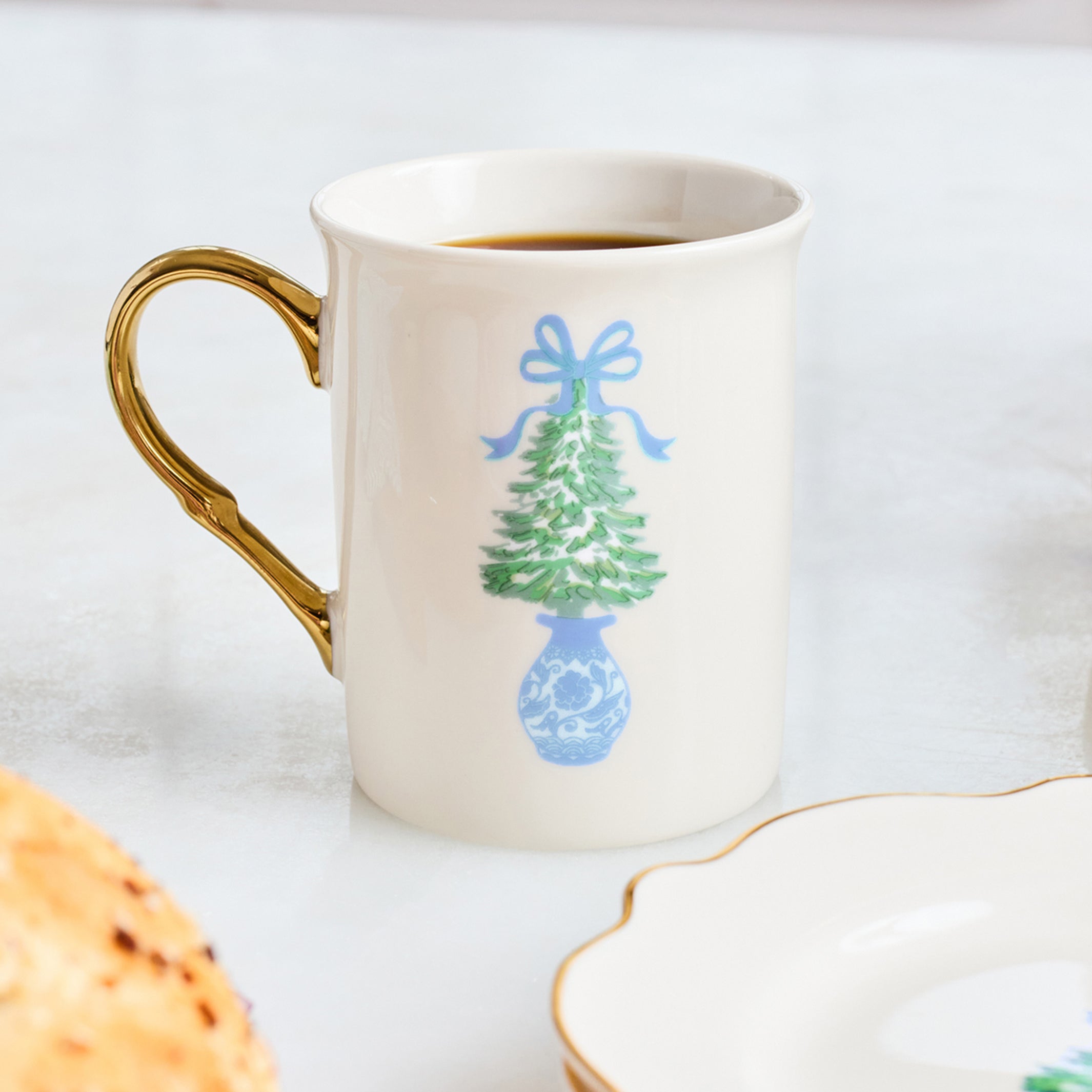 A white ceramic mug with a gold handle, filled with coffee, decorated with a blue and green Christmas tree in a blue vase. The mug is set on a light marble surface next to a plate and a pastry.