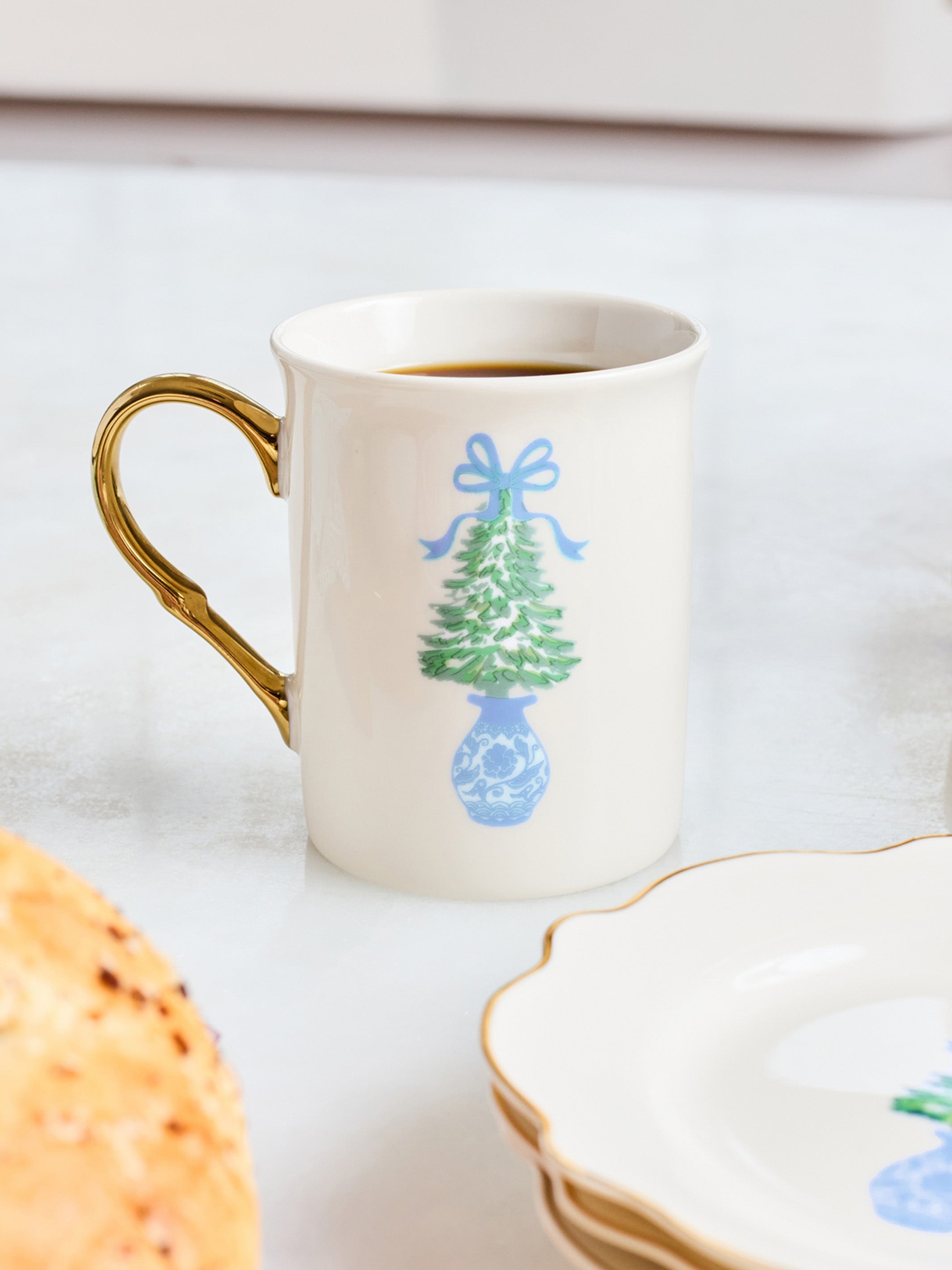 A white ceramic mug with a gold handle, filled with coffee, decorated with a blue and green Christmas tree in a blue vase. The mug is set on a light marble surface next to a plate and a pastry.