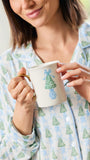 A woman in light blue and green pajamas holds a white ceramic mug with a green Christmas tree design and a gold handle.