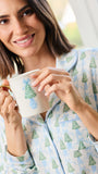 A woman wearing light blue and white pajamas with a green Christmas tree pattern holds a white ceramic mug with a gold handle in a bright indoor setting.