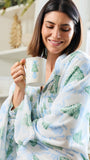 A woman wrapped in a soft white blanket with a green and light blue Christmas tree pattern, holding a white mug with a green tree design in a bright indoor setting.