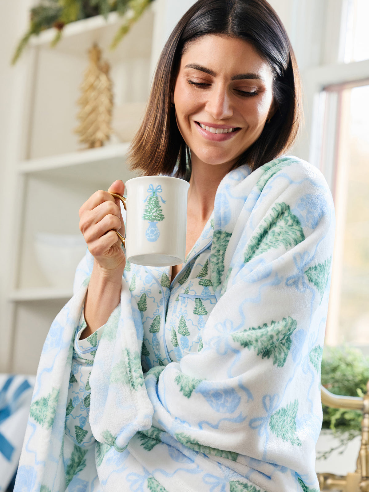 A woman wrapped in a soft white blanket with a green and light blue Christmas tree pattern, holding a white mug with a green tree design in a bright indoor setting.