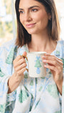 A woman in a cozy white robe with a blue and green Christmas tree pattern holds a white ceramic mug decorated with a blue bow and tree design.