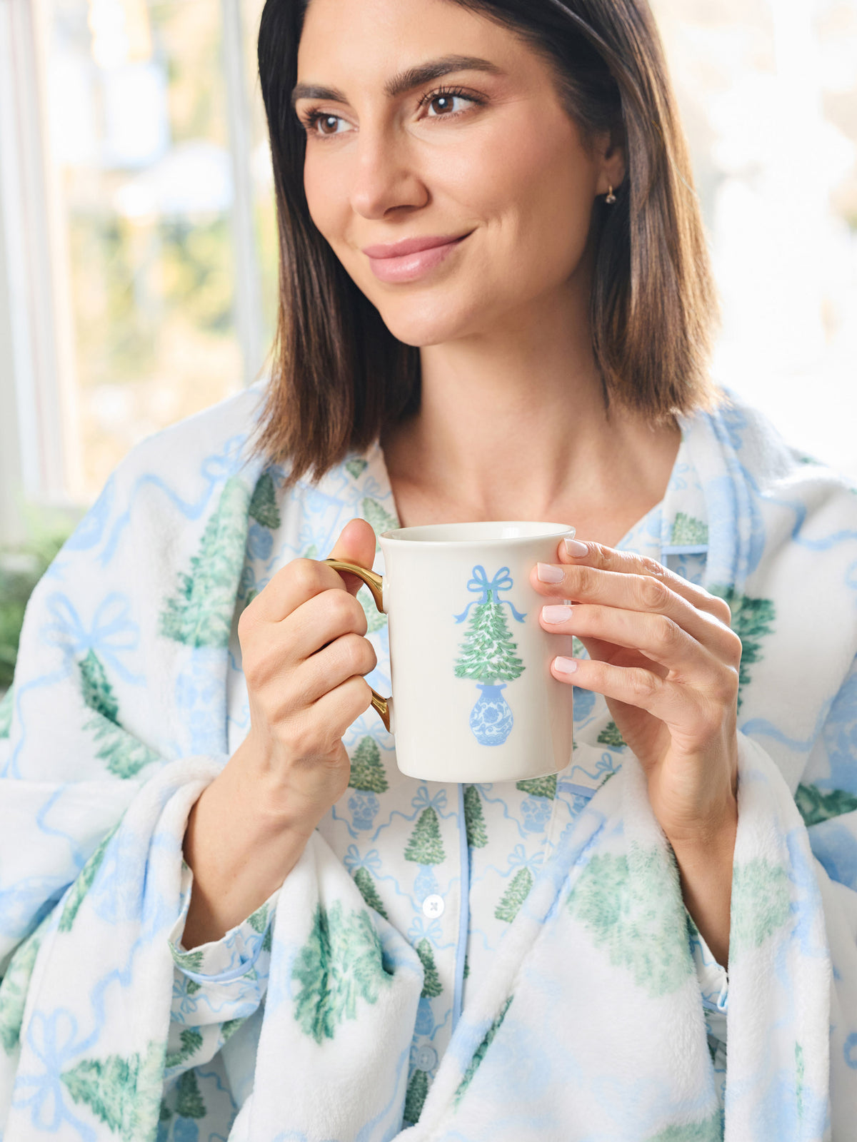 A woman in a cozy white robe with a blue and green Christmas tree pattern holds a white ceramic mug decorated with a blue bow and tree design.