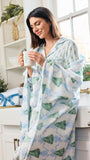 A woman in a bright kitchen wearing a white robe with a green and blue Christmas tree pattern, holding a mug.